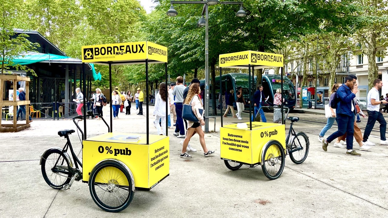 Les équipes Velo Expo pour la Mairie de Bordeaux. Distribution à vélo de Bordeaux Mag, le magazine de la Ville de Bordeaux. Photo prise place de la Comédie à Bordeaux. Campagne communication ooh hors-media par activation affichage mobile avec vélo triporteur publicitaire street marketing | Velo Expo | Agence Veymont