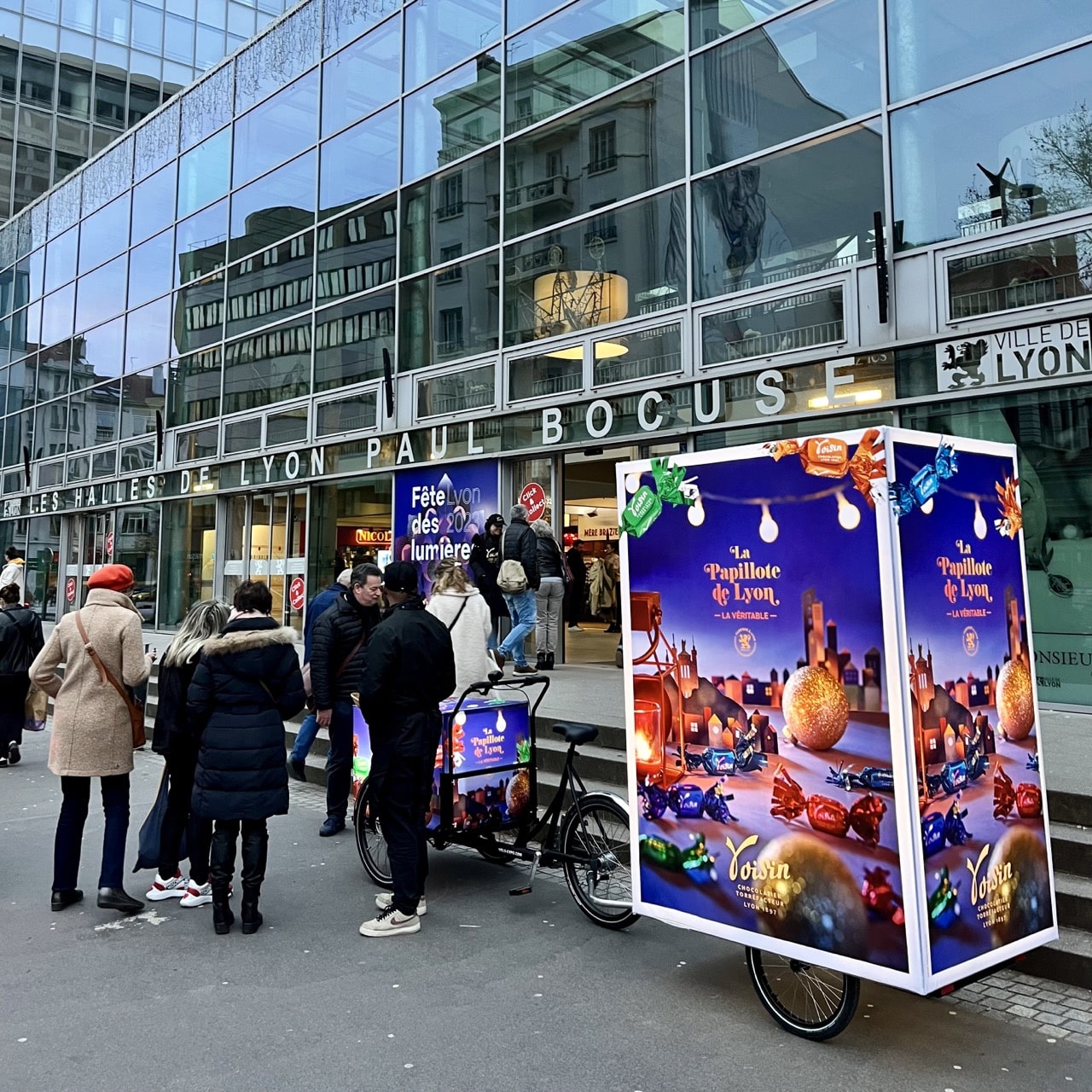 Velo Expo pour Les Chocolats Voisin, devant les Halles de Lyon Paul Bocuse. Les Papilottes Voisin. Animation, distribution, échantillonnage et dégustation avec vélo triporteur personnalisé et oriflamme publicitaire. Campagne communication ooh hors-media par activation affichage mobile avec vélo triporteur publicitaire street marketing | Velo Expo | Agence Veymont