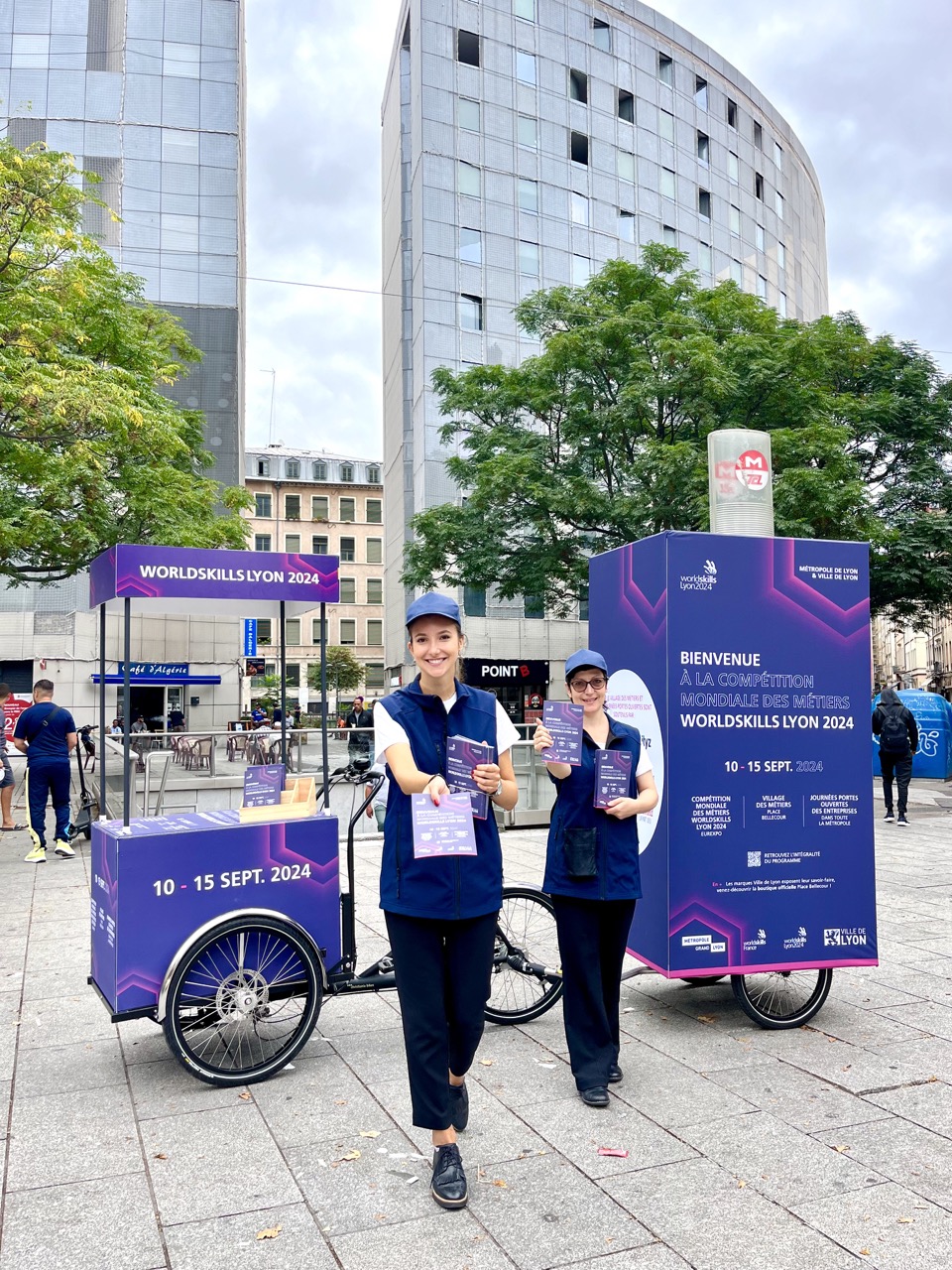 VELO EXPO x VILLE DE LYON pour la promotion de l’événement Worldskills 2024 . Photo prise à Lyon Guillotière . Animation, distribution de flyers et programmes avec vélo affichage mobile grand format. Distribution digital des programmes via QR code. Campagne communication ooh hors-media par activation affichage mobile digital avec vélo triporteur publicitaire street marketing | Velo Expo | Agence Veymont