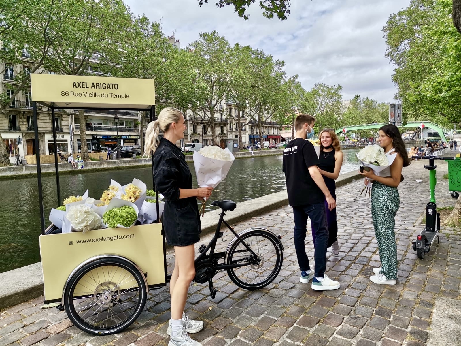 Velo Expo pour Axel Arigato Paris Marais. Affichage mobile, animation drive-to-store, distribution de fleurs avec vélo triporteur corgo personnalisé. Campagne communication ooh hors-media par activation affichage mobile digital avec vélo triporteur publicitaire street marketing | Velo Expo | Agence Veymont